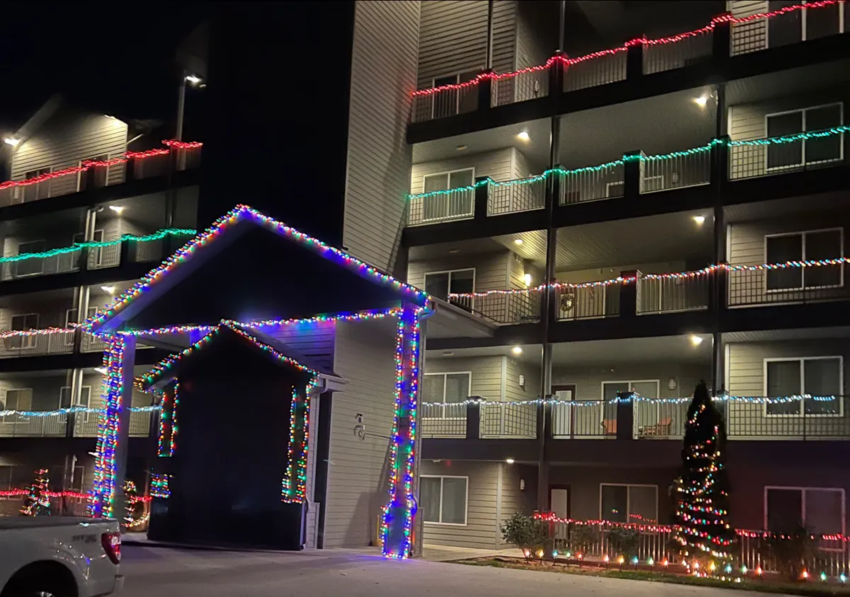 A festive exterior scene featuring decorative lights on the building, a grand entrance structure, and a twinkling Christmas tree, evoking celebration and togetherness.