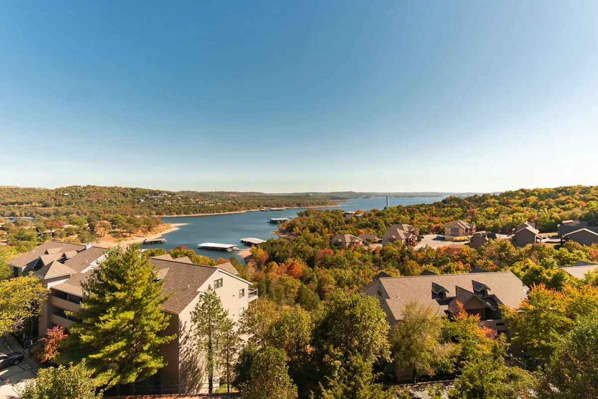 This image captures a sweeping view of Table Rock Lake with mountains in the background, brilliant autumn foliage, and residential buildings nestled by the shoreline. The tranquil water invites boating, fishing, and other recreational pursuits, while the scenery is framed by natural beauty and vibrant colors.