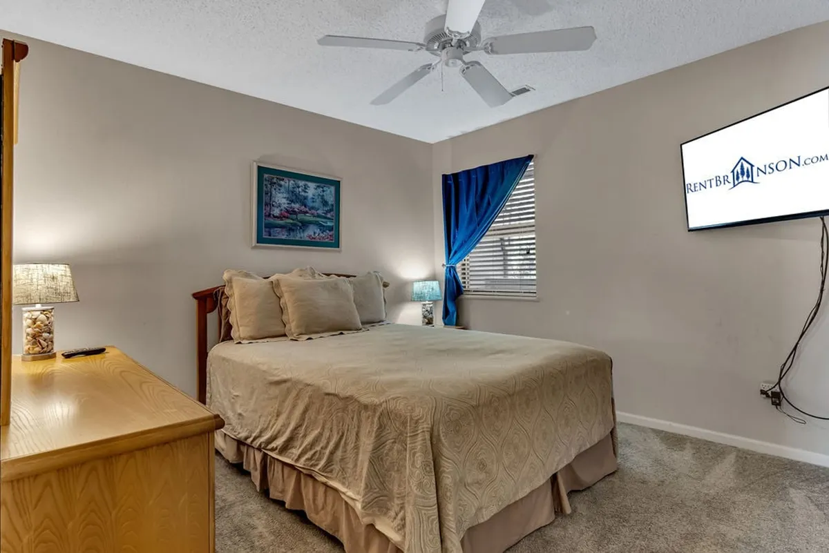 A beautifully appointed bedroom featuring a plush bed with crisp linens, a nightstand with a lamp for evening reading, a flat-screen television, and a large window with soft curtains that allow natural light to fill the room.