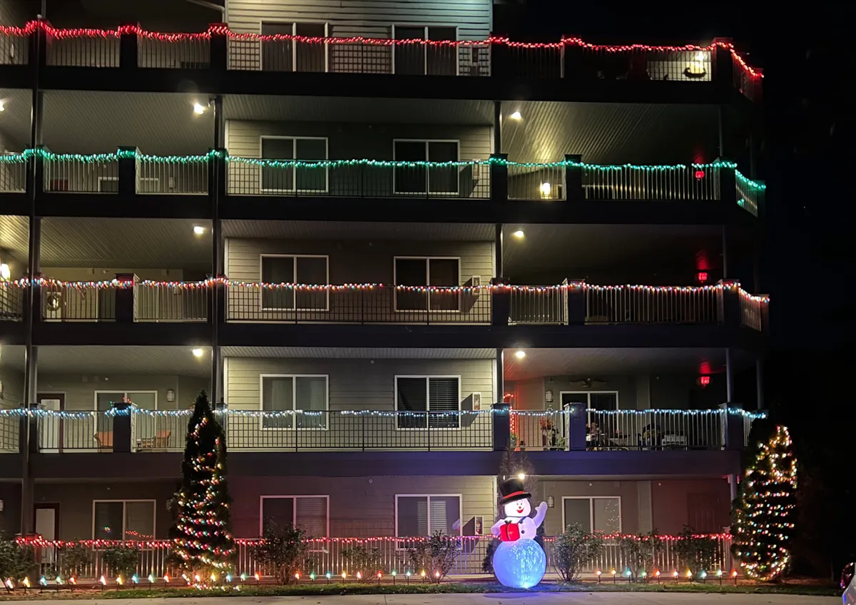 An exterior shot featuring festive Christmas lights, a snowman decoration, and a Christmas tree outside the condo, set against a backdrop that hints at a community environment. The scene conveys a sense of celebration, community, and outdoor enjoyment.
