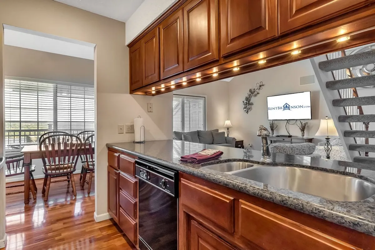 The image showcases a seamless, open-plan interior featuring a granite countertop kitchen with wooden cabinets that flows directly into a spacious dining area and a cozy living room with inviting seating. The overall ambiance is warm, ideal for both cooking and gathering, and provides a clear visual of how the spaces are interconnected for group activities.