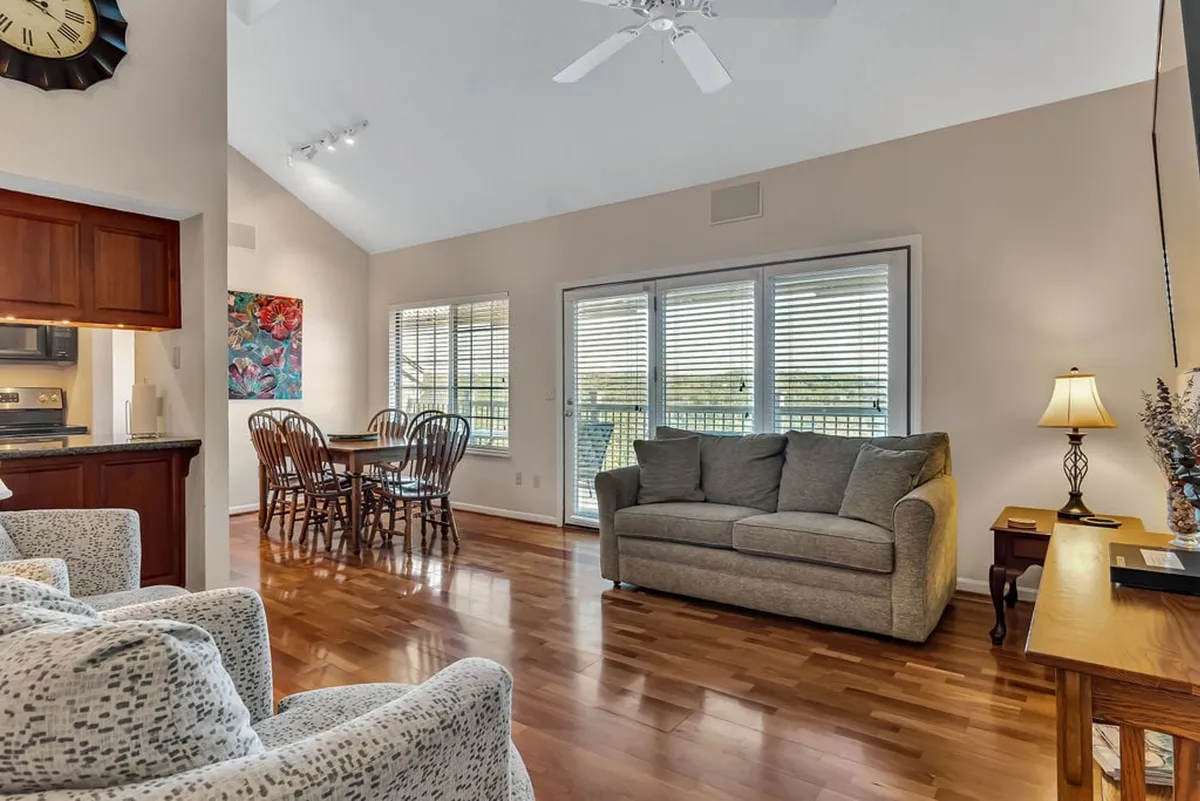 A bright and inviting interior space with a comfortable sofa, a dining table set near large windows, and artwork on the walls. The large windows hint at views to the outdoors and allow natural light to pour in, creating a seamless transition from the interior to the exterior experience.