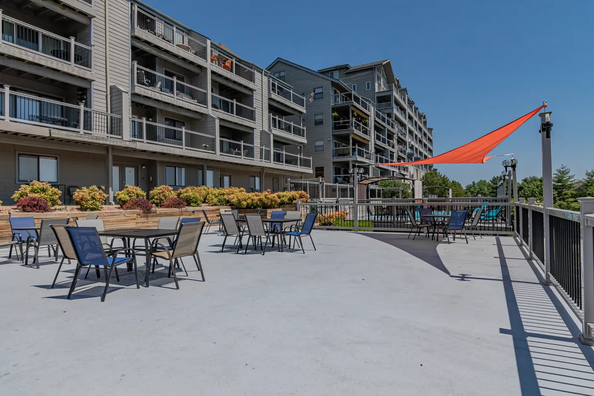 The image features a cheerful outdoor seating area beneath a vibrant orange shade sail, bordered by lush flower beds and overlooked by condo balconies. The communal setup encourages social gatherings or relaxing outdoor moments.