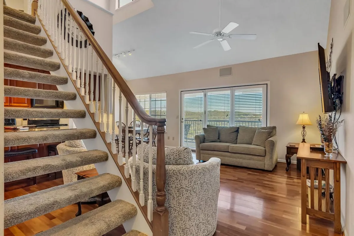 An open-plan living space with a sofa, dining table, large windows, and a view of the staircase, integrating relaxation, dining, and socializing. The abundance of natural light and the flow between seating and dining areas support a wide range of group activities and gatherings.