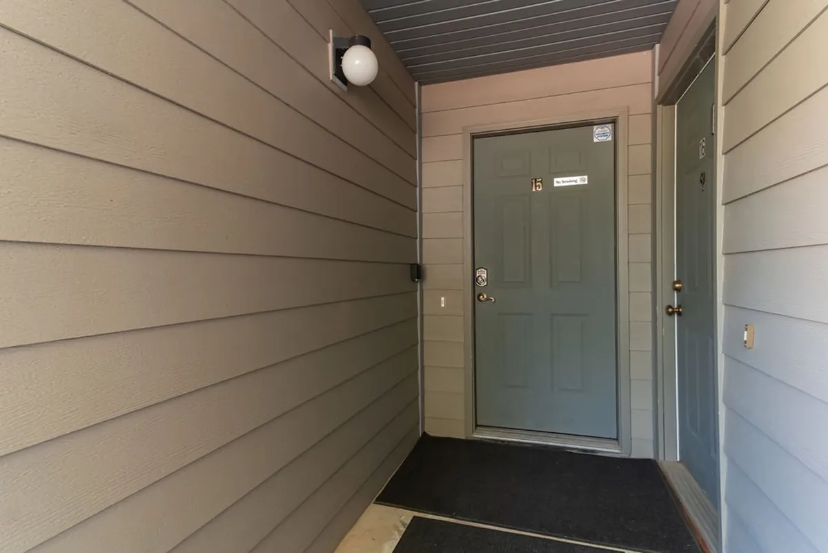 The main entrance door of the condo, with a clean doormat and a warm light fixture above. The entryway is tidy and inviting, suggesting a sense of arrival and welcome.